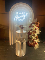 Captivating engagement party ambiance with stylish backdrops, exquisite floral arrangements, an intricately designed cake, and the warm glow of neon lights spelling out 'it was always you