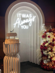 Engagement party scene featuring decorative backdrops, lush floral arrangements, a beautifully designed cake, and neon lights with the phrase 'it was always you